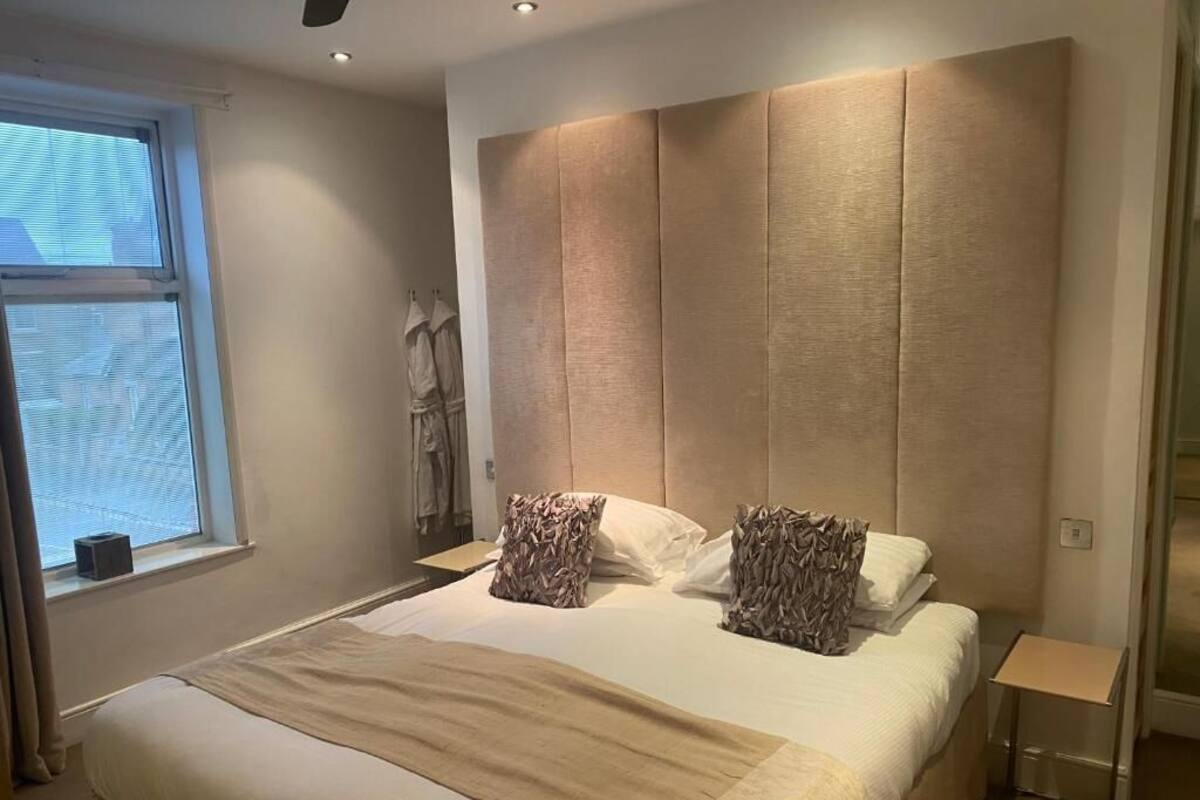 Un dormitorio con una cama grande, cabecera, ventana y mesita de noche.