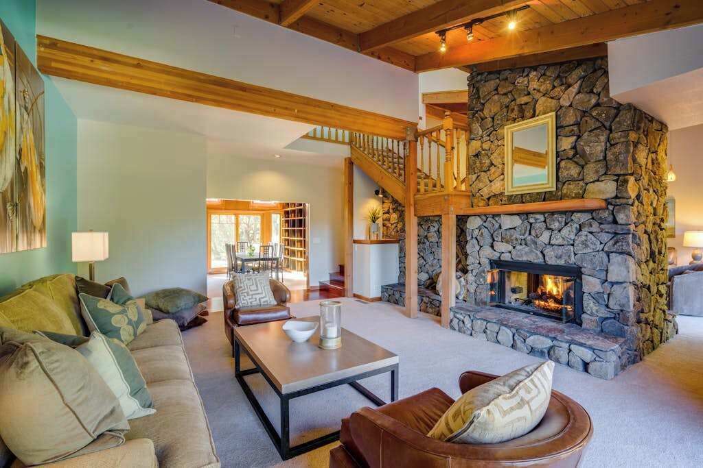 A cozy living room with a stone fireplace, wooden beams, a staircase, and a dining area.