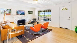 A living room with a fireplace, orange chairs, a sofa, and a view of greenery outside. - Healdsburg 3BR vineyard farmhouse with game barn & wood fireplace (Healdsburg)