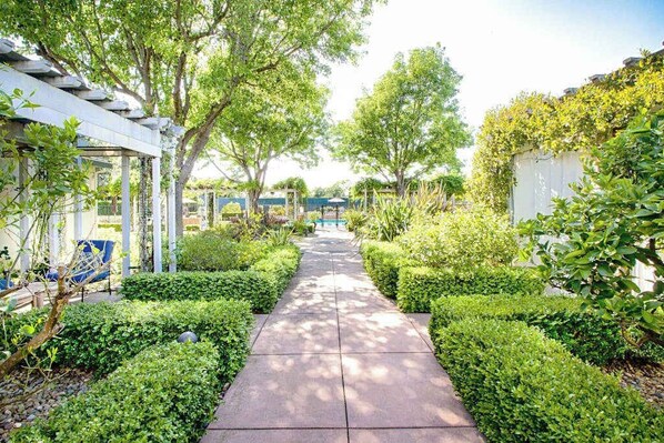Un sendero bordeado por setos y árboles bien cuidados, que lleva a una zona cubierta con una pérgola.