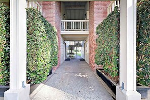 A covered walkway flanked by brick walls and green hedges, leading to a gated entrance.