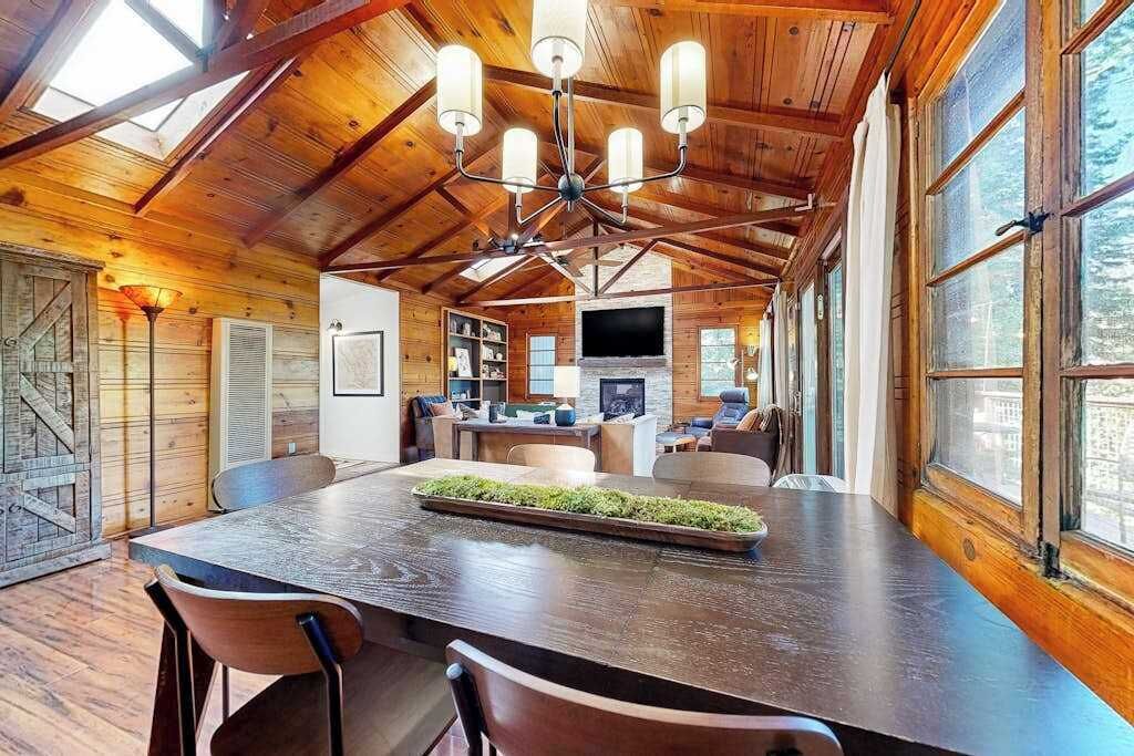 A spacious wooden cabin interior with a large dining table, multiple chairs, a fireplace, and large windows.