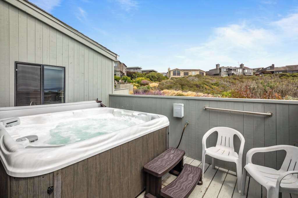 A hot tub on a deck with a railing, two chairs, and a view of houses in the background.