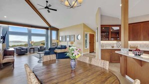A spacious living area with a large wooden dining table, comfortable seating, a kitchen with wooden cabinets, and large windows offering a view. - Bodega Harbour Ocean-View Hot Tub Retreat (Bodega Bay)