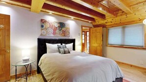Ein Schlafzimmer mit einem Bett, einem runden Tisch mit Lampe, einem farbenfrohen abstrakten Gemälde und Holzbalkendecke.