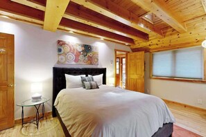A bedroom with a bed, a round table with a lamp, a colorful abstract painting, and wooden ceiling beams. - Monte Rio 2BR A-frame cabin with private hot tub — dog friendly (Monte Rio)