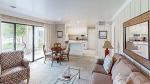 A living room with a sofa, coffee table, and dining area with chairs. There is a kitchen in the background with white cabinets and a dining table.