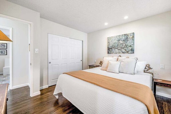 A bedroom with a large bed, a white door, a nightstand, and a wall-mounted artwork.