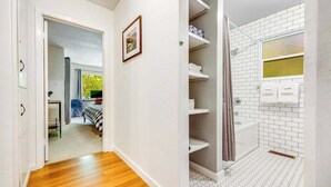 Een gang die leidt naar een slaapkamer met een bed, een badkamer met een douche en een betegelde vloer.