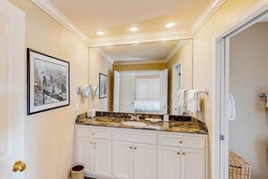 Une salle de bain avec un meuble-lavabo, un évier et un miroir. Des serviettes sont accrochées au mur et un panier est posé sur le sol.