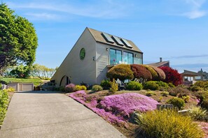 A house with a sloped roof, large windows, and a well-maintained garden with various plants and flowers. - Bodega Bay 2BR ocean-view home with pool table, on the golf course (Bodega Bay)