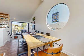 A dining area with a long table, chairs, a bench, a round window, and a view of the outdoors. - Bodega Bay 2BR ocean-view home with pool table, on the golf course (Bodega Bay)