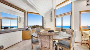 A dining area with a round table, wicker chairs, and large windows with ocean views. - Bodega Bay 5BR ocean-view home with hot tub — dogs welcome (Bodega Bay)