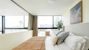 Una camera d'hotel con un'ampia finestra, un letto con lenzuola bianche, una coperta lavorata a maglia marrone e un quadro raffigurante nuvole sulla parete.