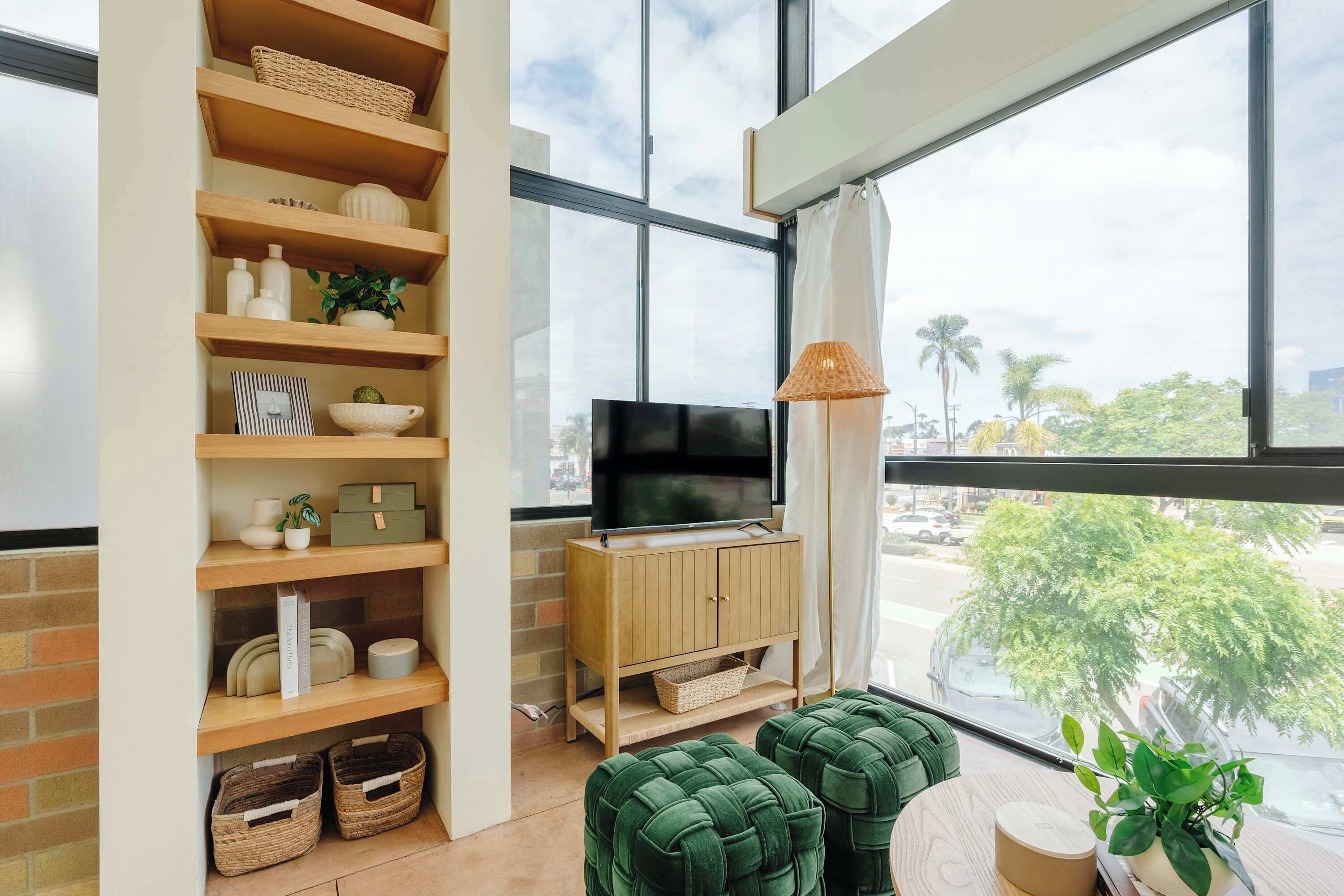 A modern living room with a TV, wooden shelves, and a large window with a view of palm trees.