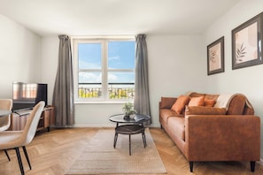 A living room with a sofa, TV, dining table, and chairs, a small table with a plant, and a view through a window.