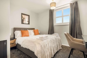 Una habitación de hotel con cama, una silla, una ventana con cortinas, una mesita de noche y una lámpara fijada a la pared.