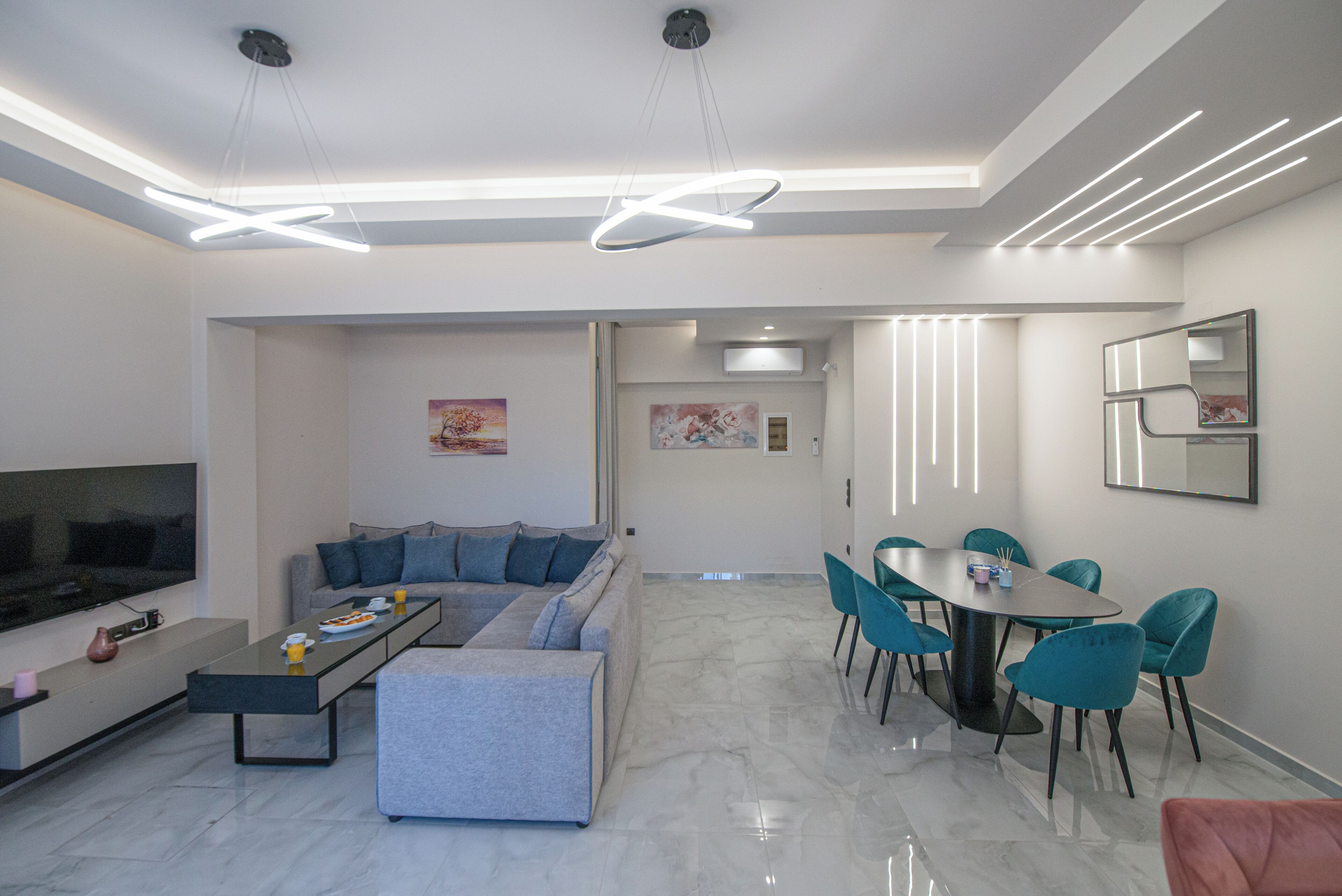A modern living room with a sectional sofa, a flat-screen TV, a dining area with a table and chairs, and a kitchen in the background.