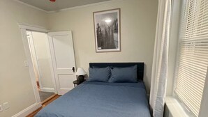 Ein Schlafzimmer mit einem Bett, einem gerahmten Bild an der Wand, einem Fenster mit Jalousien und einer Tür.