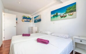 Une chambre à coucher avec un lit, deux oreillers, une table de chevet, une étagère murale ornée de chapeaux et un tableau représentant une scène de plage.