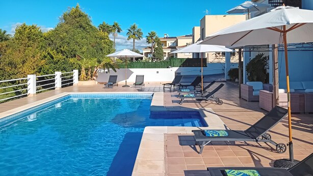 Una piscina con sillas de descanso y sombrillas, rodeada por una valla blanca y palmeras.