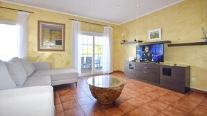 Una sala de estar con un sofá, una mesa de centro de vidrio, un televisor sobre un mueble bajo y un ventanal corredero que da a un espacio exterior.