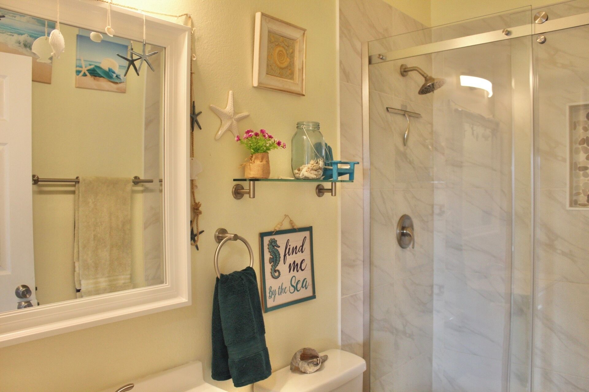 Une salle de bain dotée d’un grand miroir, d’un porte-serviettes, d’une douche et d’une étagère où sont disposés des objets décoratifs.