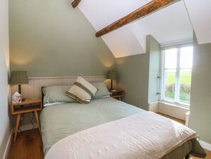 Ein Schlafzimmer mit geneigter Decke, einem Bett mit gesteppter Bettdecke, zwei Nachttischlampen und einem Fenster mit Blick auf Grünflächen.