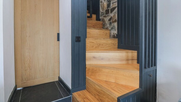 Una escalera de madera junto a un muro de piedra, una puerta de madera y un suelo negro.