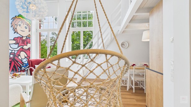 Una habitación con una silla colgante de cuerda, una zona de comedor con sillas blancas y un ventanal con vistas a árboles.