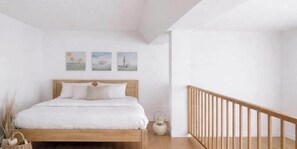 Ein Schlafzimmer mit einem hölzernen Bett, weißer Bettwäsche, einem kleinen Nachttisch und einem Beistelltisch. An der Wand hängen drei Bilder von Segelbooten.