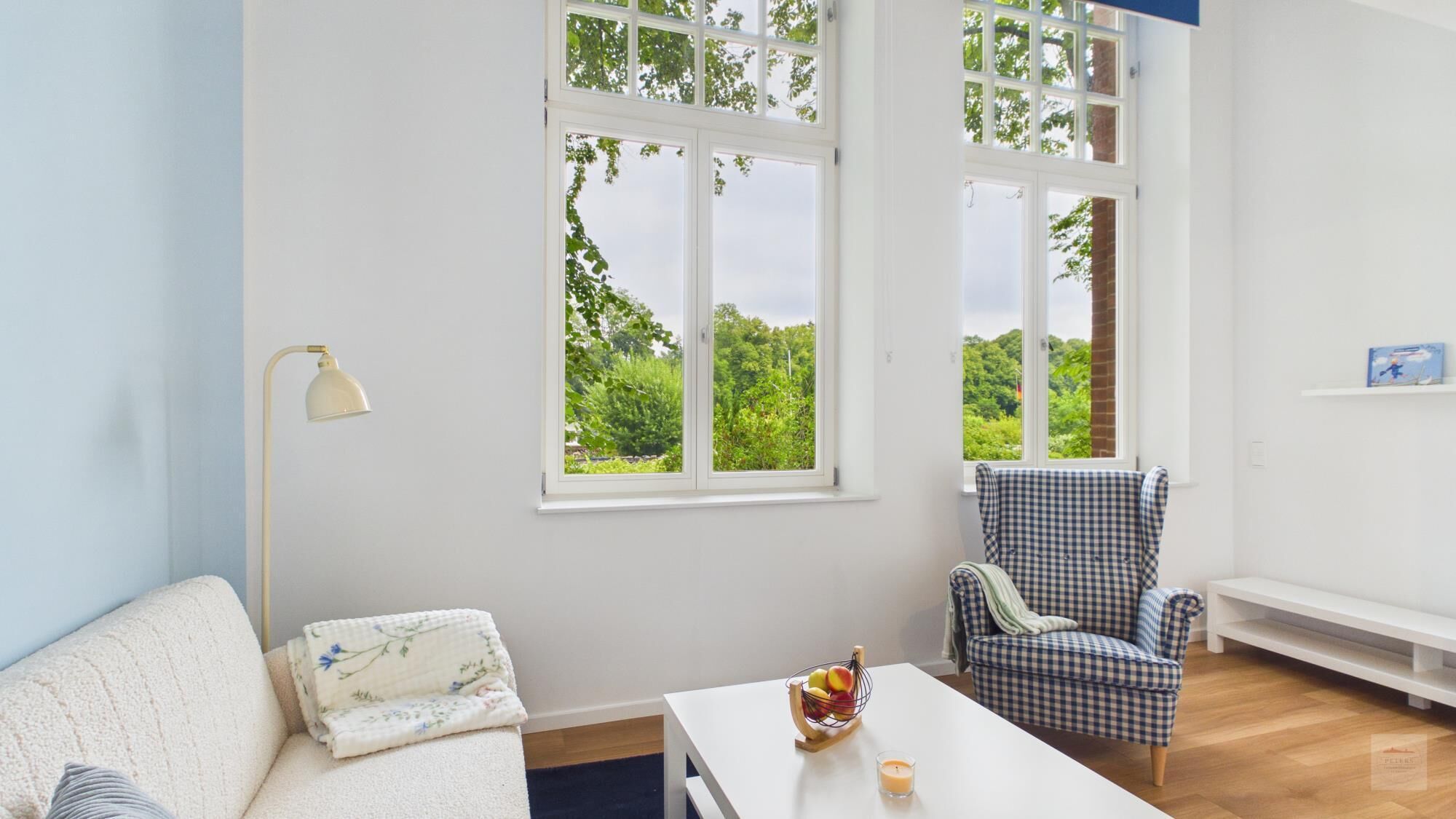 Een kamer met een witte bank, een zwart-wit gestreepte fauteuil, een kleine tafel met een schaal fruit en een kaars, en grote ramen met uitzicht op groen.