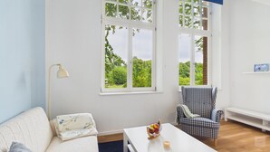 Habitación con sofá blanco, sillón a cuadros, una pequeña mesa con frutero y vela, y ventanas grandes con vistas a zonas verdes.