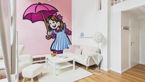 Une pièce avec un canapé blanc, une table basse blanche, un fauteuil blanc moelleux, un mur rose orné d’une illustration d’une fillette tenant un parapluie, et une lampe blanche.
