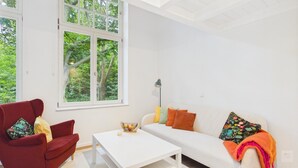Una sala de estar moderna con un sofá rojo, una mesa de centro blanca y un sofá blanco con cojines coloridos. Amplias ventanas ofrecen vistas a árboles al exterior.