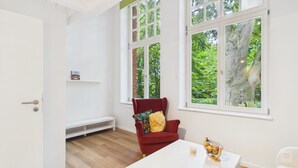 Una habitación con una silla roja, una mesa blanca y ventanas amplias con vistas a árboles.