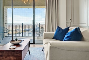Ein Wohnzimmer mit einer Couch, einem Couchtisch mit einer Schale und einem Buch, sowie einem Blick durch eine Glastür auf einen Außenbalkon mit Stühlen und einem Tisch.