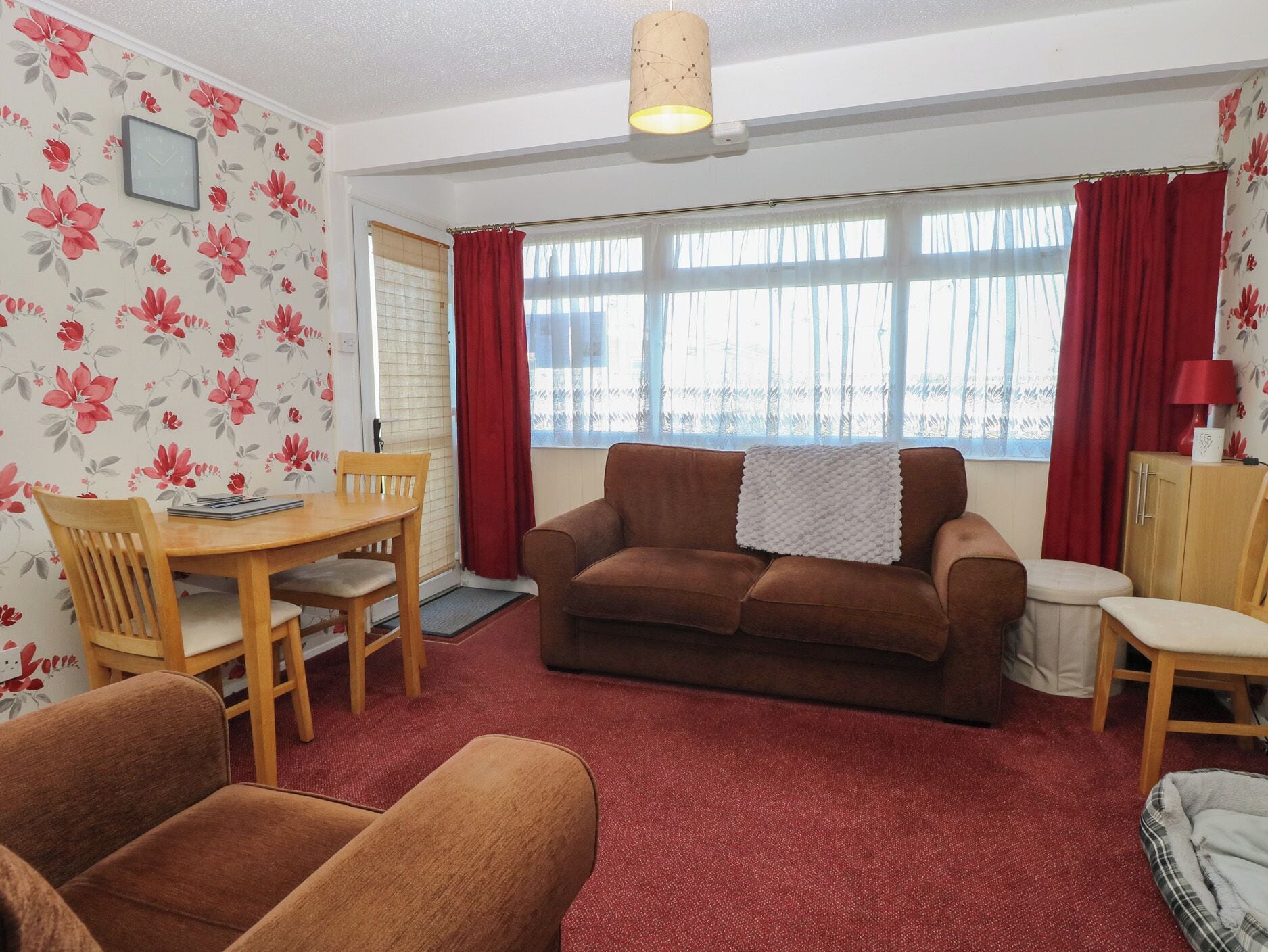 A living room with a brown sofa, wooden dining table, and chairs, a red carpet, and a window with red curtains.