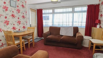 A living room with a brown sofa, wooden dining table, and chairs, a red carpet, and a window with red curtains.