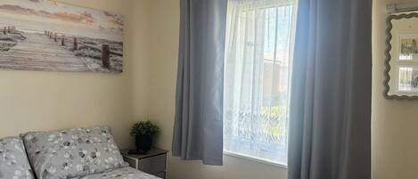 Un dormitorio con cama, mesita de noche, planta en maceta, ventana con cortinas y un cuadro en la pared.