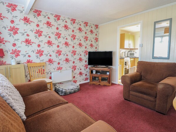 A living room with a red carpet, floral wallpaper, a television, and a kitchen visible through an open door. - Mon Chalet (Great Yarmouth)