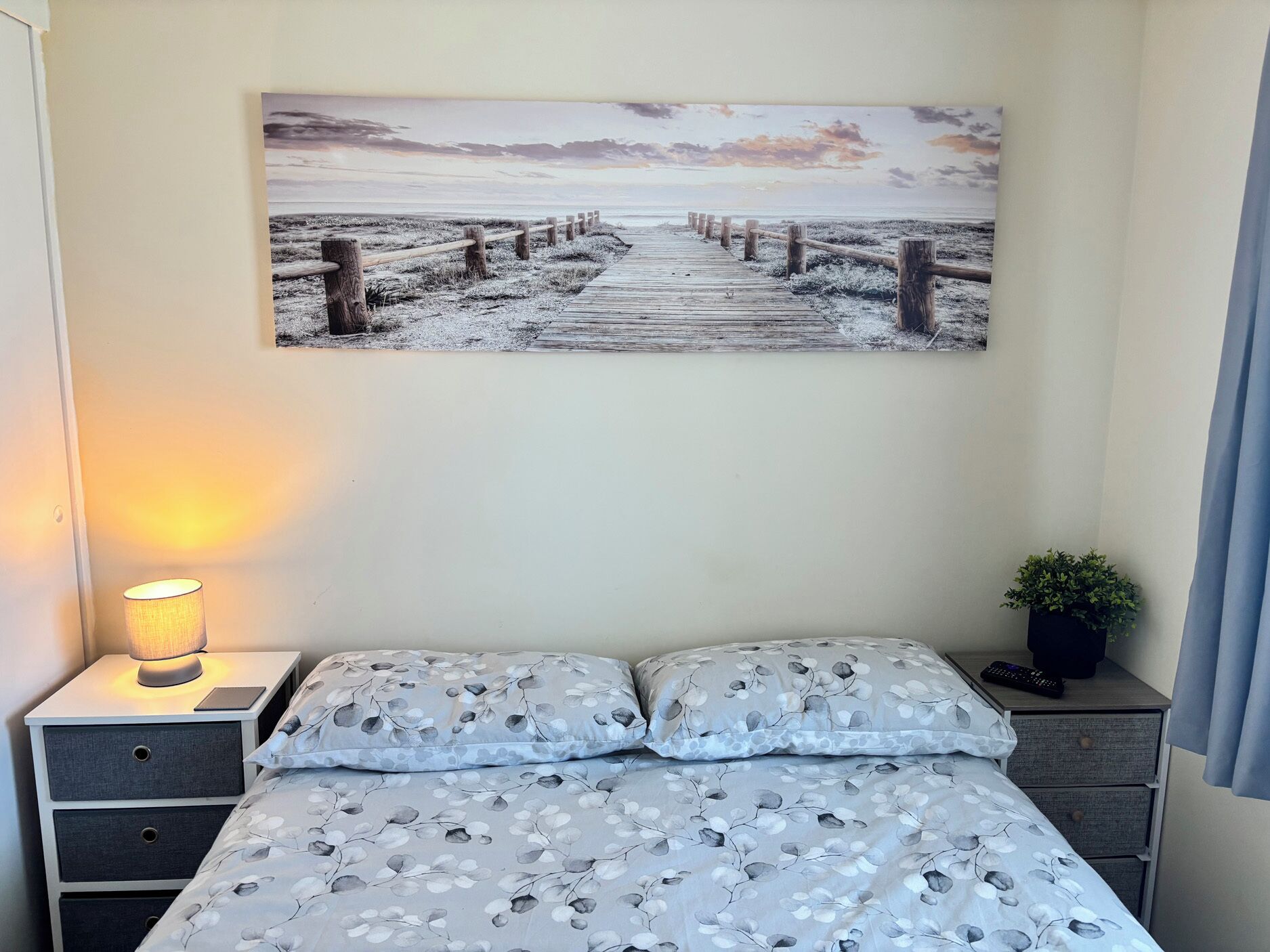 Quarto com cama, mesa de cabeceira com abajur, quadro na parede e um vaso pequeno com planta.
