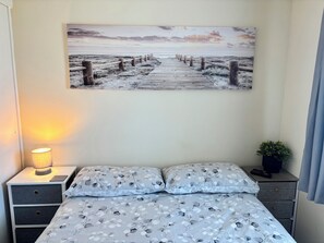 A bedroom with a bed, a nightstand with a lamp, a wall-mounted painting, and a small potted plant.