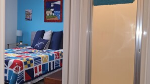 Un dormitorio con cama, mesita de noche, lámpara, ventana con vistas y puerta con una toalla colgada.