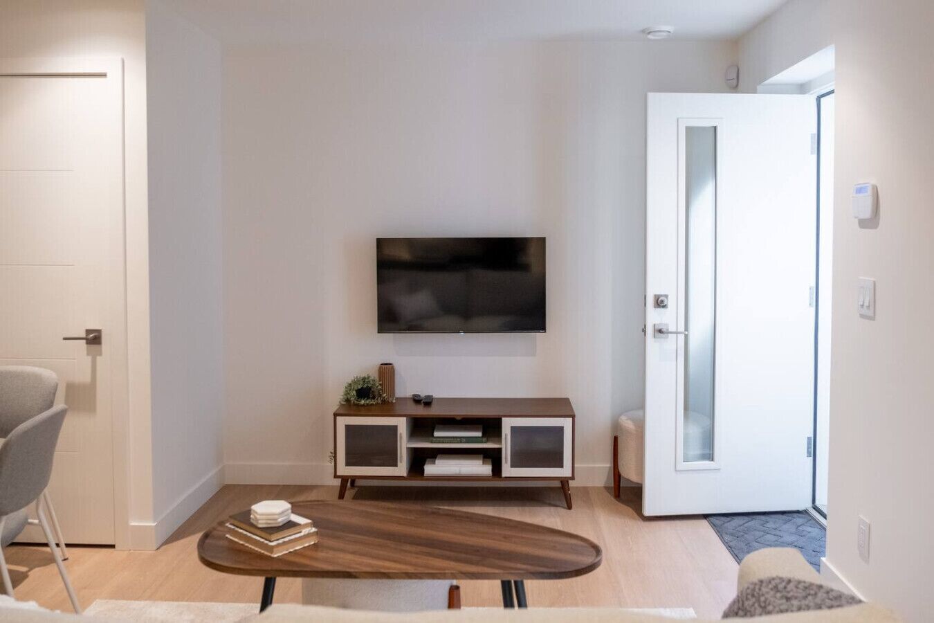 A modern living room with a flat-screen TV mounted on the wall, a wooden coffee table, a grey armchair, and a white door.