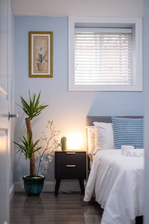 A bedroom with a bed, bedside table, lamp, window, and a potted plant. - Walk to SkyTrain, Grocery & More 2 bedroom suite (Vancouver)