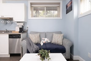 A compact living space with a kitchenette, a grey sofa with patterned cushions, a small table with a plant, and a window with blinds. - Walk to SkyTrain, Grocery & More 2 bedroom suite (Vancouver)