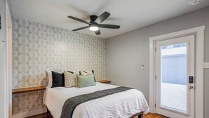 A bedroom with a bed, a ceiling fan, a patterned wallpaper, a door with a view, and a wooden floor. - Urban Retreat Near Downtown Houston with Gated Access (Houston)