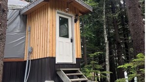 Una piccola baita di legno con una porta bianca, un piccolo portico e terreno sterrato.