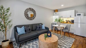 Un salon moderne avec un canapé gris, une table basse ronde en bois et un coin cuisine doté de meubles blancs et d’un réfrigérateur blanc.